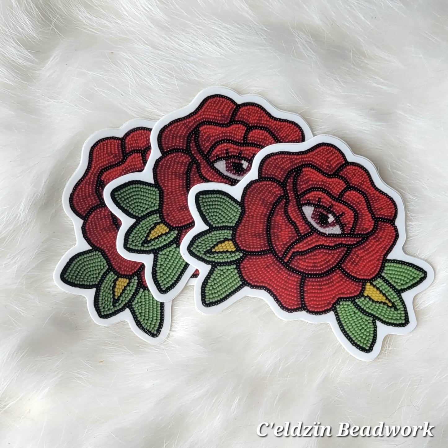 A sticker of a beaded red rose patch with green leaves on each side and a brown eye in the center.