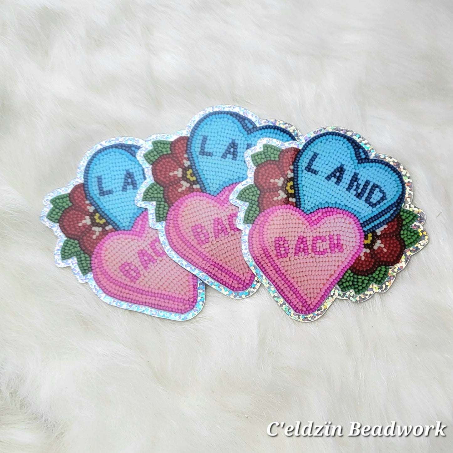 A sticker of a beaded Land Back statement piece. It is two candy hearts, one blue and one pink, with the blue one on top and the pink one on the bottom. The blue says Land the pink says Back. There's one red flower on each side of it with green leaves. It also has a glittery border.