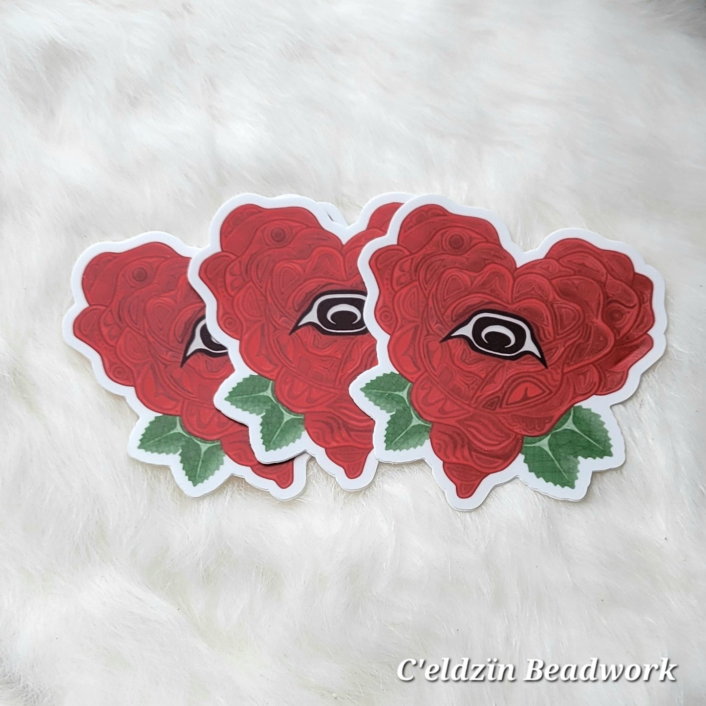 A sticker of a red heart-shaped rose with formline art in each petal. The center has a formline styled brown eye, and it has two green leaves on each side on the bottom half.