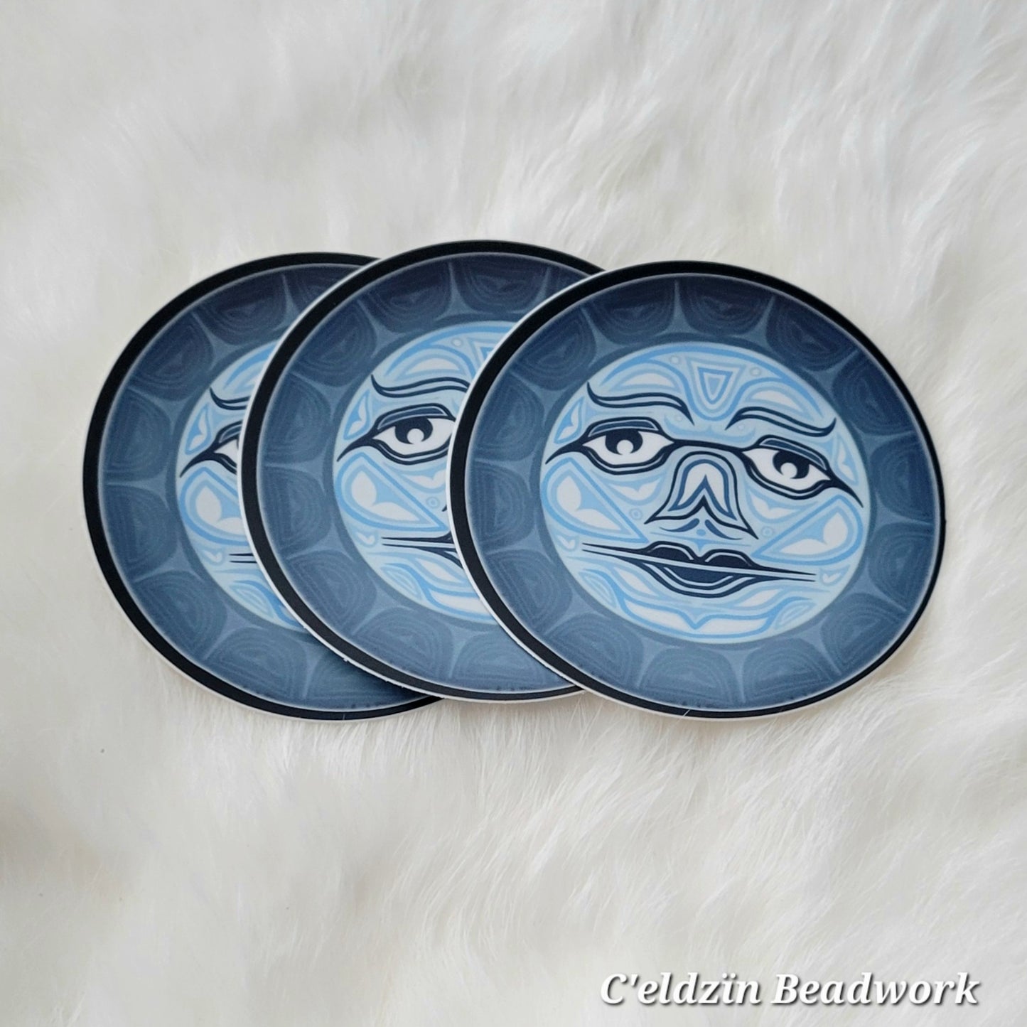A sticker of a formline styled moon with a face. It is a light blue moon with white filled formline shapes. It has a thick dark blue outline.