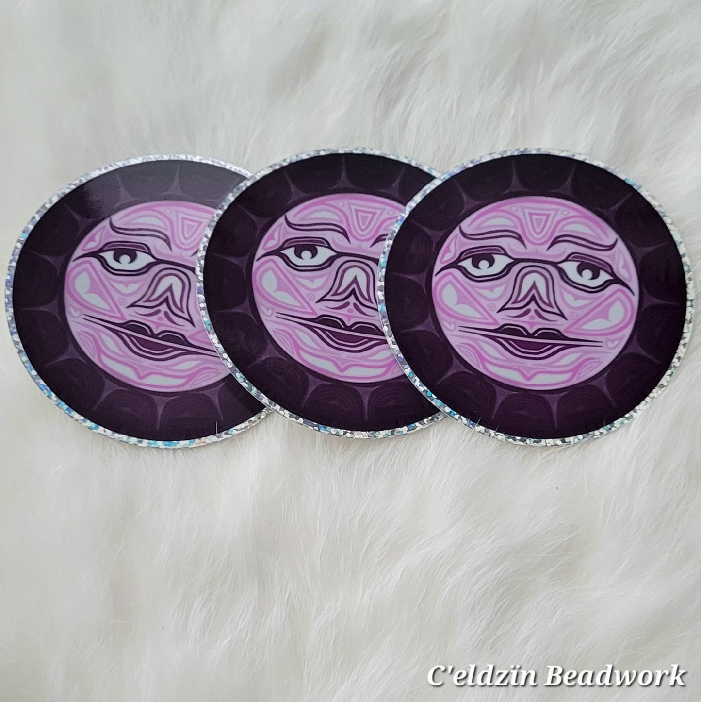 A sticker of a formline styled moon with a face. It is pink with white filled formline shapes and a thick dark purple-pink outline.