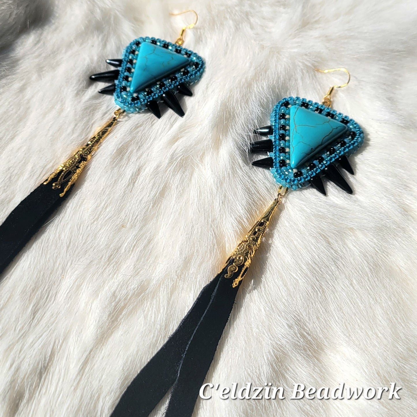 Upside down, triangle shaped turquoise howlite stone with turquoise, white, and black beads embroidered around it and one row of turquoise rhinestone banding. It has three black spikes on each side in order of small, big, then small. It hangs on gold sterling silver hooks and has a gold cone attached to the bottom with black dyed deer hide fringe.
