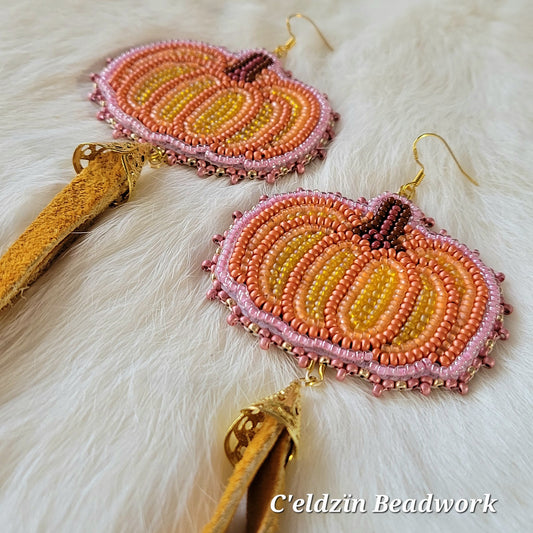 Bead embroidered orange pumpkin earrings with a brown stem and pink outline. It hangs on gold sterling silver hooks and has a gold cone with two strings of tanned moose hide. 